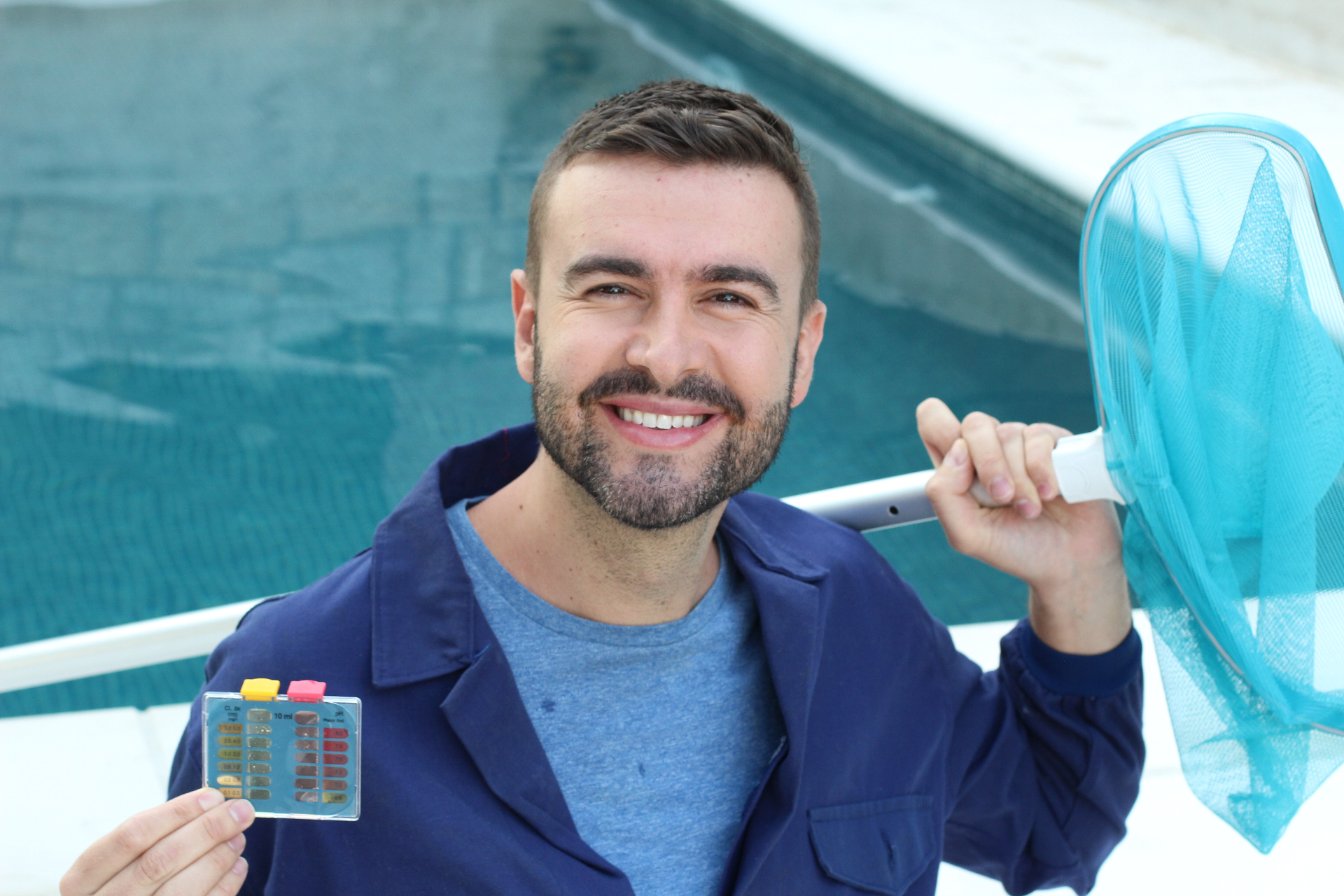 Smiling pool technician holding a water test kit and skimmer net by a pool